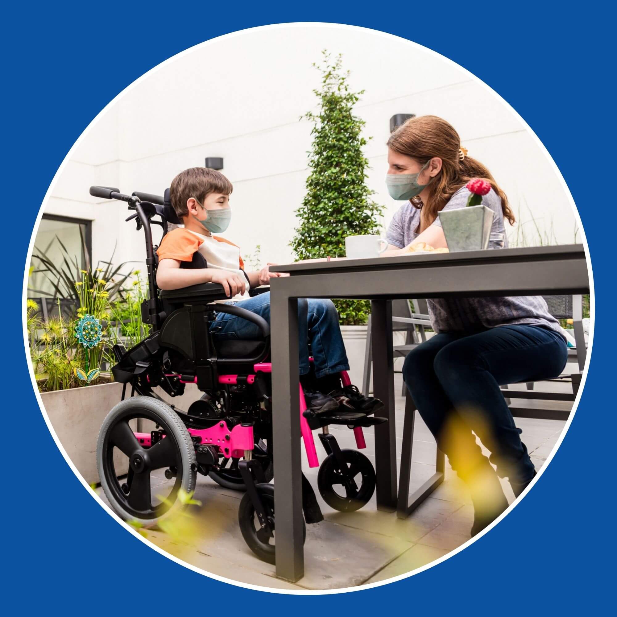 The photo shows a young boy in a wheelchair sitting at a table in a garden with a young woman. Image is from iStock-1280050807