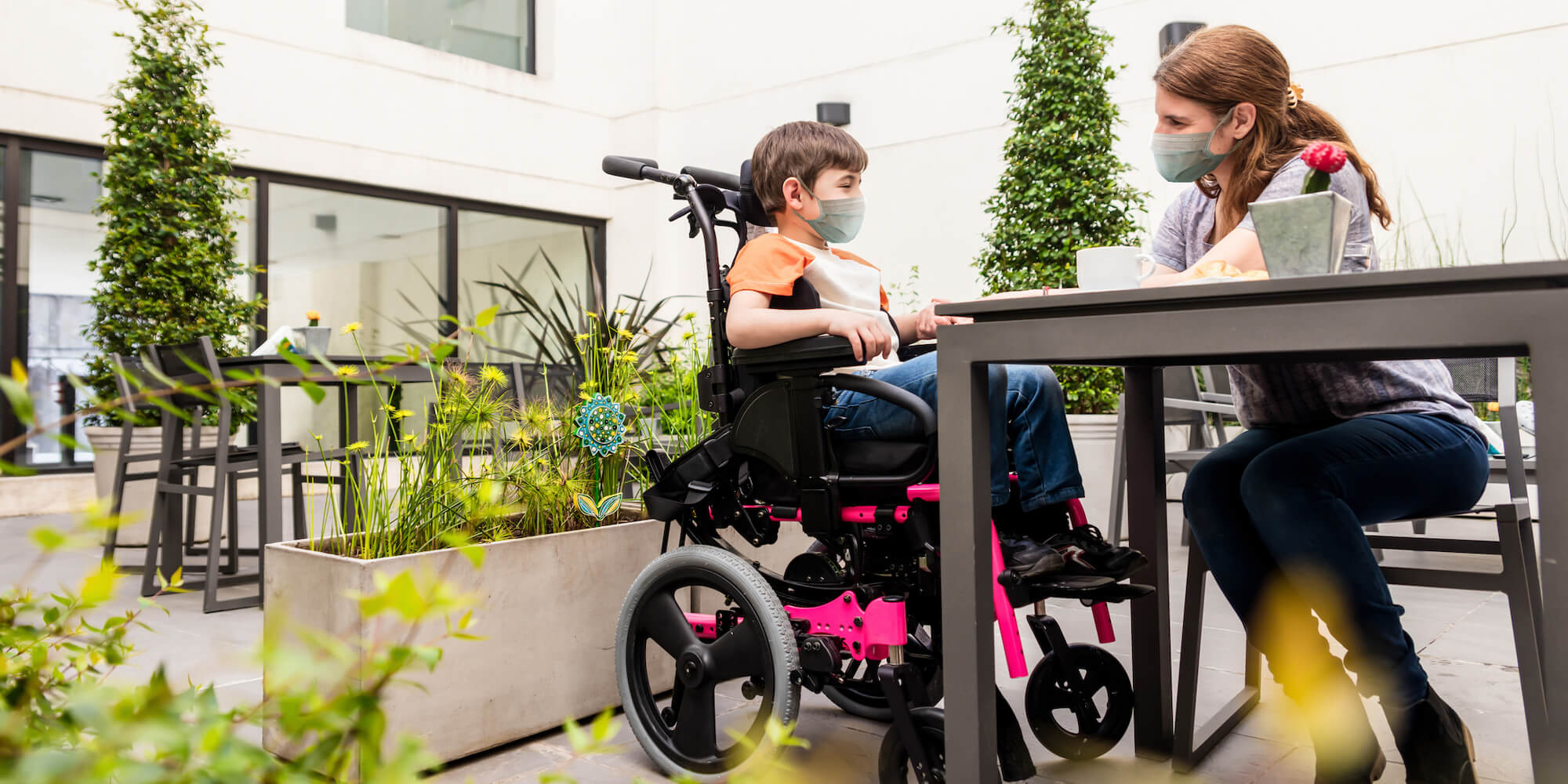 The photo shows a young boy in a wheelchair sitting at a table in a garden with a young woman. Image is from iStock-1280050807 